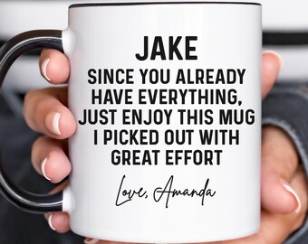 Puede incluir: Taza de cerámica blanca con asa y borde negros. La taza tiene el texto "JAKE SINCE YOU ALREADY HAVE EVERYTHING, JUST ENJOY THIS MUG I PICKED OUT WITH GREAT EFFORT Love, Amanda." La taza es sostenida por una persona.
