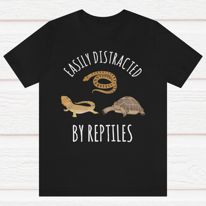 May include: Black t-shirt with white text that reads "Easily Distracted By Reptiles". The shirt features a cartoon image of a snake, a lizard, and a turtle.