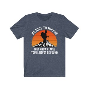 Op de afbeelding: Een marineblauw T-shirt met een witte afbeelding van een wandelaar met een rugzak en wandelstokken voor een zonsondergang. De tekst luidt "Wees aardig tegen wandelaars, ze kennen plekken waar je nooit gevonden zult worden."