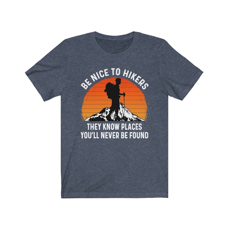 Op de afbeelding: Een marineblauw T-shirt met een witte afbeelding van een wandelaar met een rugzak en wandelstokken voor een zonsondergang. De tekst luidt "Wees aardig tegen wandelaars, ze kennen plekken waar je nooit gevonden zult worden."