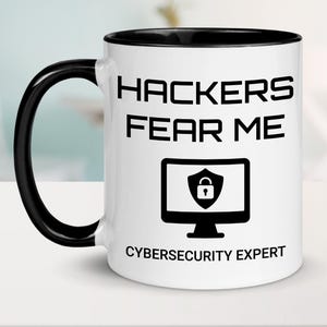 May include: White coffee mug with a black handle and black rim. The mug has black text that reads "Hackers Fear Me Cybersecurity Expert" and a black icon of a computer monitor with a padlock inside.