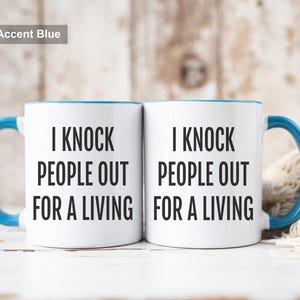 May include: Two white ceramic mugs with blue accents, each featuring the text "I KNOCK PEOPLE OUT FOR A LIVING" in black. The mugs have blue handles and rims. The text "11oz Accent Blue" is in the top left corner.