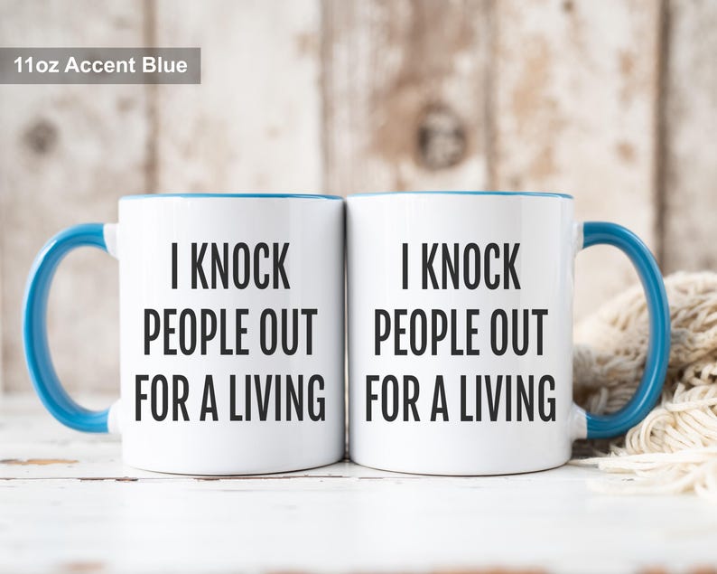 May include: Two white ceramic mugs with blue accents, each featuring the text "I KNOCK PEOPLE OUT FOR A LIVING" in black. The mugs have blue handles and rims. The text "11oz Accent Blue" is in the top left corner.