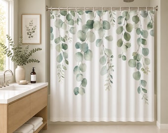 Op de afbeelding: Een wit douchegordijn met een aquarelprint van hangende eucalyptus-takken in diverse tinten groen en salie. Het botanische patroon loopt vanaf de bovenkant naar beneden en hangt aan metalen ringen aan een zilverkleurige roede.