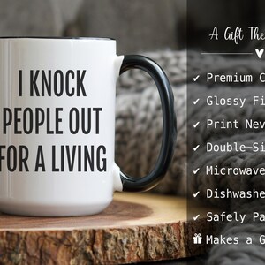May include: White ceramic mug with a black handle and rim. The mug features the text "I KNOCK PEOPLE OUT FOR A LIVING" in bold black letters. The mug is on a wooden surface.