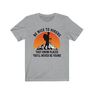 Op de afbeelding: Grijs T-shirt met een silhouet van een wandelaar voor een zonsondergang. De tekst luidt "Wees aardig tegen wandelaars, ze kennen plekken waar je nooit gevonden zult worden."