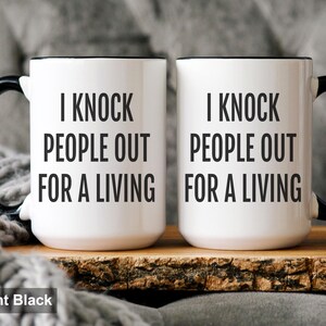 May include: Two white ceramic mugs with black handles and rims. The mugs have the text "I KNOCK PEOPLE OUT FOR A LIVING" printed in black. The mugs are displayed on a wooden surface.