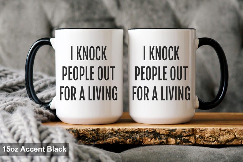 May include: Two white ceramic mugs with black handles and rims. The mugs have the text "I KNOCK PEOPLE OUT FOR A LIVING" printed in black. The mugs are displayed on a wooden surface.
