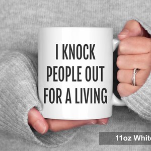 May include: White ceramic mug with the text "I KNOCK PEOPLE OUT FOR A LIVING" in black. The mug is held by a person wearing a gray sweater. The mug is labeled "11oz White".