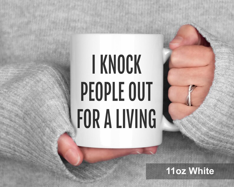 May include: White ceramic mug with the text "I KNOCK PEOPLE OUT FOR A LIVING" in black. The mug is held by a person wearing a gray sweater. The mug is labeled "11oz White".