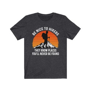 Op de afbeelding: Een donker grijs gem&ecirc;leerd t-shirt met een witte afbeelding van een wandelaar met een rugzak en een bergketen op de achtergrond. De tekst "Be Nice To Hikers They Know Places You'll Never Be Found" staat op het t-shirt gedrukt.