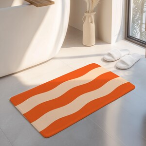 May include: A rectangular bath mat featuring a repeating pattern of wide, wavy horizontal stripes in alternating shades of vibrant orange and soft cream. The mat has rounded corners and a smooth, flat surface texture throughout the design.