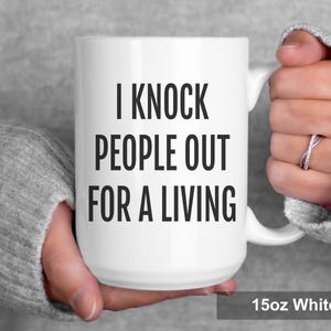 May include: White ceramic mug with the text "I Knock People Out For A Living" in bold black letters. The mug is 15oz and has a white handle. The mug is held by a person wearing a gray sweater.