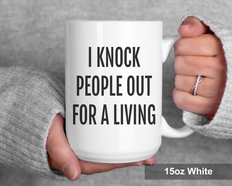 May include: White ceramic mug with the text "I Knock People Out For A Living" in bold black letters. The mug is 15oz and has a white handle. The mug is held by a person wearing a gray sweater.