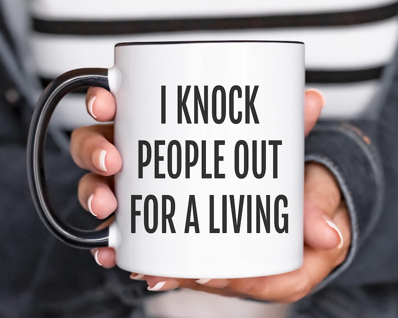 May include: White ceramic mug with black interior and handle. The mug features the text "I KNOCK PEOPLE OUT FOR A LIVING" in bold black letters. The mug is held by a person.