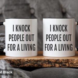 May include: Two white coffee mugs with black handles and rims. The mugs have the text "I KNOCK PEOPLE OUT FOR A LIVING" printed in black. The mugs are 11oz in size and are displayed on a wooden surface.