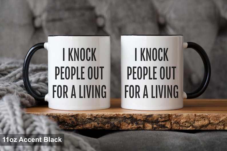 May include: Two white coffee mugs with black handles and rims. The mugs have the text "I KNOCK PEOPLE OUT FOR A LIVING" printed in black. The mugs are 11oz in size and are displayed on a wooden surface.