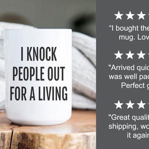 May include: White ceramic mug with the text "I KNOCK PEOPLE OUT FOR A LIVING" in black. The mug has a large handle and is displayed on a wooden surface. Customer reviews are visible in the background.