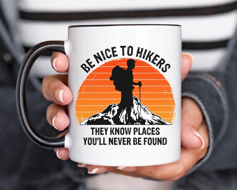 May include: White ceramic mug with a black handle. The mug features a graphic of a hiker with a backpack and a sunset in the background. The text on the mug reads "Be Nice To Hikers They Know Places You'll Never Be Found."