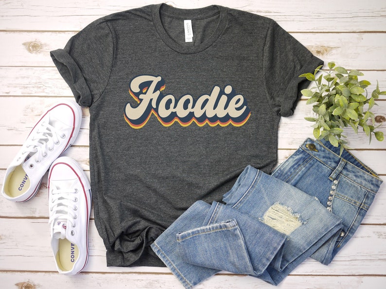 Puede incluir: Una camiseta gris oscuro con la palabra "Foodie" impresa en una fuente de estilo retro con acentos naranja, amarillo y azul. La camiseta est&aacute; colocada sobre una superficie de madera blanca con un par de zapatillas blancas y unos vaqueros azules.