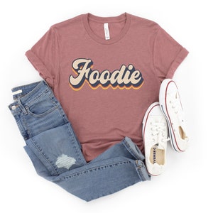 Puede incluir: Una camiseta color malva con la palabra "Foodie" en una fuente retro y colorida. La camiseta se combina con jeans azules y zapatillas blancas.