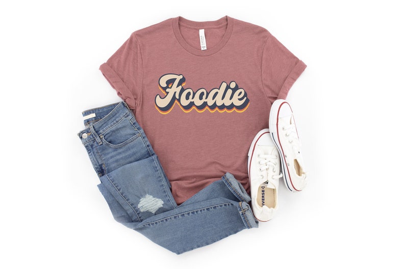 Puede incluir: Una camiseta color malva con la palabra "Foodie" en una fuente retro y colorida. La camiseta se combina con jeans azules y zapatillas blancas.
