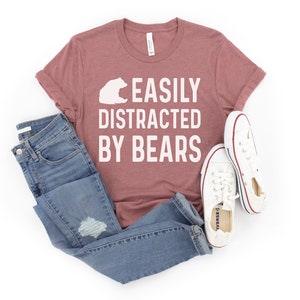 May include: A mauve t-shirt with the text "EASILY DISTRACTED BY BEARS" in white lettering. A white bear silhouette is above the text. The t-shirt is paired with blue jeans and white sneakers.