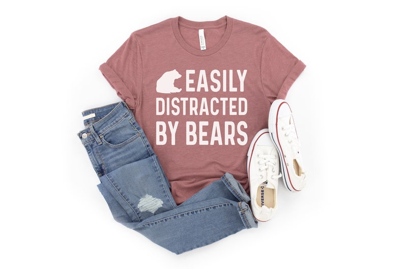May include: A mauve t-shirt with the text "EASILY DISTRACTED BY BEARS" in white lettering. A white bear silhouette is above the text. The t-shirt is paired with blue jeans and white sneakers.