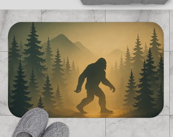 Puede incluir: Una alfombrilla de baño rectangular con la silueta oscura de una criatura Bigfoot caminando a través de un denso bosque de pinos frente a un paisaje montañoso brumoso de color amarillo dorado, con tonos tierra.