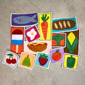 May include: A collection of colorful felt food patches, including a fish, carrot, bread, ice pop, fried egg, hot dog, broccoli, corn, cherries, pear, strawberry, orange, and bell pepper. Each patch is square with a decorative border.
