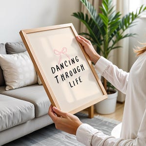 Puede incluir: Un cuadro enmarcado con las palabras "DANCING THROUGH LIFE" en letras negras. Un lazo rosa está encima del texto. El marco es de color marrón claro. El cuadro se sostiene frente a un sofá gris y una planta verde.