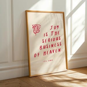 Op de afbeelding: Een ingelijste print met de quote "JOY IS THE SERIOUS BUSINESS OF HEAVEN" in rode letters. Een rood bloemmotief staat in de linkerbovenhoek. De lijst is lichtbruin van kleur en de achtergrond is gebroken wit.