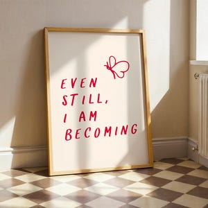 Puede incluir: Un cartel enmarcado con las palabras "EVEN STILL, I AM BECOMING" en escritura roja. Un gráfico de mariposa roja está encima del texto. El marco es de color marrón claro y el fondo es blanquecino. El cartel está apoyado contra una pared blanca.