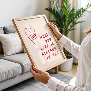 May include: A framed print with a light beige background and a red heart-shaped design. The text "WHAT YOU LOVE IS ALREADY IN YOU" is written in red. The frame is made of light-coloured wood.