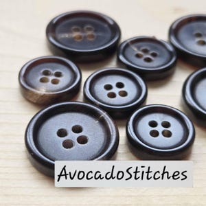 May include: A collection of dark brown, circular buttons with four holes in the center. Each button features a smooth, polished surface and a raised outer rim. The text "AvocadoStitches" is printed in black on a white rectangular label.
