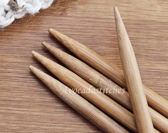 Könnte beinhalten: Ein Set aus fünf hellbraunen Stricknadeln aus Holz, die fächerförmig angeordnet sind. Die Nadeln haben glatte, spitz zulaufende Enden und eine sichtbare Holzmaserung. Ein Teil eines weißen gehäkelten Textils ist am oberen Rand zu sehen.