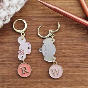 May include: Two gold stitch markers with clear animal charms and pink and orange letter charms. The charms are shaped like a bunny and an elephant. The letter charms say "R" and "W".