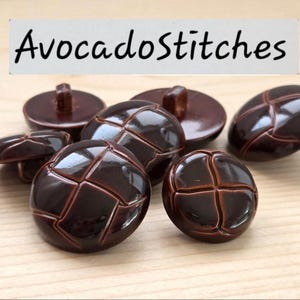 May include: A collection of round, dark brown buttons with a glossy finish and a cross-shaped embossed pattern on the surface. The text "AvocadoStitches" is displayed in black lettering at the top of the image above the buttons.