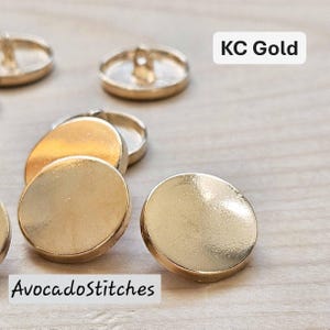 May include: Close-up of several KC Gold buttons with a shiny, metallic finish. The buttons are round with a flat face and a loop on the back for sewing. The text "KC Gold" and "AvocadoStitches" are visible.