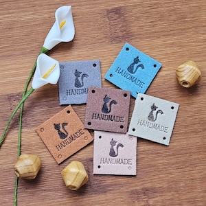 May include: Six brown leather tags with the word "HANDMADE" and a silhouette of a cat printed on them. The tags are arranged on a wooden surface.