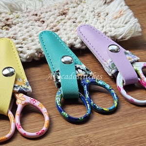 May include: Three pairs of small scissors with colourful floral patterned fabric covers and leather sheaths. The scissors have blue, green, and yellow handles and are on a wooden surface.