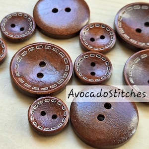 Peut inclure: Divers boutons en bois brun foncé de différentes tailles. Certains boutons ont une bordure blanche cousue décorative. Chaque bouton a deux trous pour la couture. Le texte "AvocadoStitches" est visible.