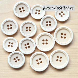 May include: A selection of off-white buttons, each with four holes. The buttons feature a light beige central area and a slightly raised, rounded rim. The text "AvocadoStitches" is visible. These buttons are suitable for sewing and craft projects.