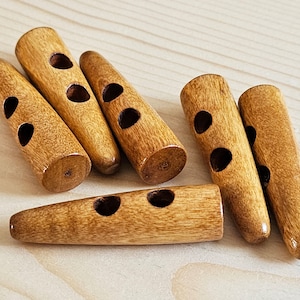 May include: A collection of six light brown wooden toggle buttons, each featuring a cylindrical, tapered shape with two circular holes drilled through the center for attachment, showcasing a natural wood grain texture and a smooth finish.