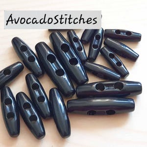 May include: A collection of black toggle buttons with a smooth, glossy finish. Each button features a long, cylindrical shape with two centered holes for attachment. The text "AvocadoStitches" is visible in the upper left corner of the image.
