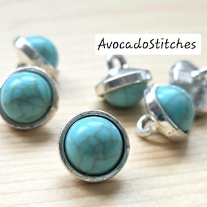 May include: Close-up of several turquoise and silver buttons. Each button features a round, light blue stone with a webbed pattern, set in a silver-colored metal casing. The text "AvocadoStitches" is in the upper right corner.