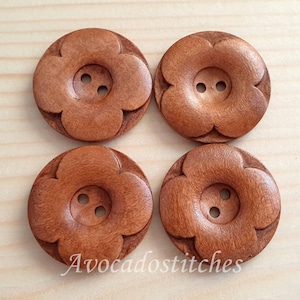May include: Four circular brown wooden buttons, each featuring a carved four-petal flower design and two central holes for attachment. The text Avocadostitches is printed in white at the bottom of the image, centred below the four buttons.
