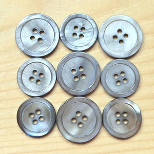 May include: A collection of nine circular buttons arranged in a three-by-three grid. Each button is made of a smooth, gray, iridescent material with a subtle sheen. Every button features four small holes in the center for attachment to fabric.