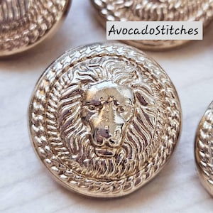 May include: Close-up of several gold-coloured metal buttons. Each button features a detailed lion's head design surrounded by a textured border. The text "AvocadoStitches" is visible in the upper right corner. These buttons are suitable for sewing and craft projects.