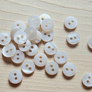 May include: A collection of white pearl buttons with two holes each. The buttons are arranged in a random pattern on a light wood surface.